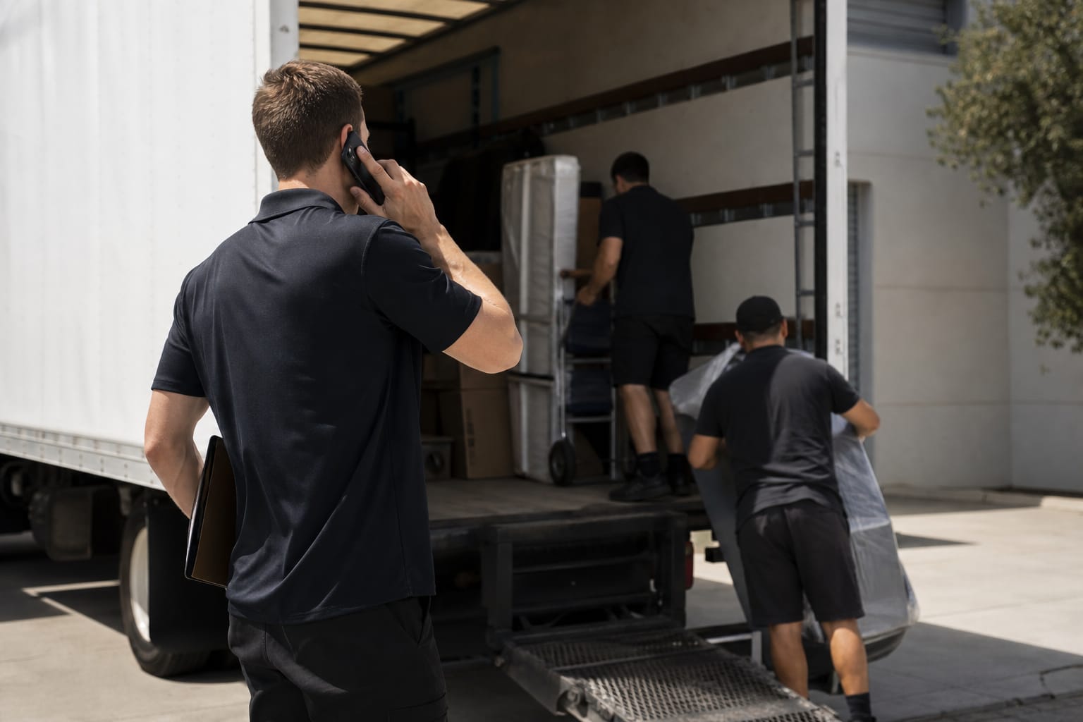Mover on the phone with a clipboard while two crew load a dolly behind him in a truck bay