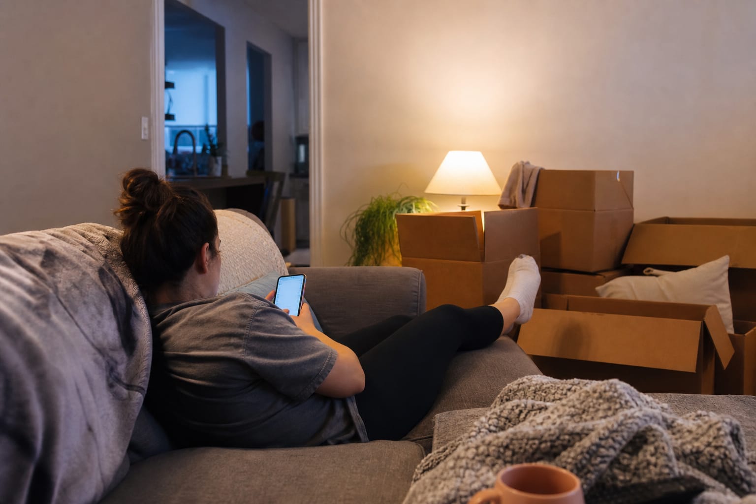 Homeowner scrolling her phone at night surrounded by half-packed cardboard moving boxes