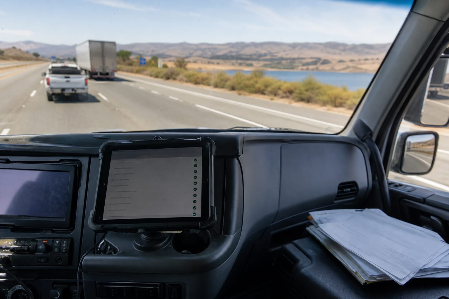Tablet on a truck dashboard showing a job checklist beside a paper bill of lading, highway ahead