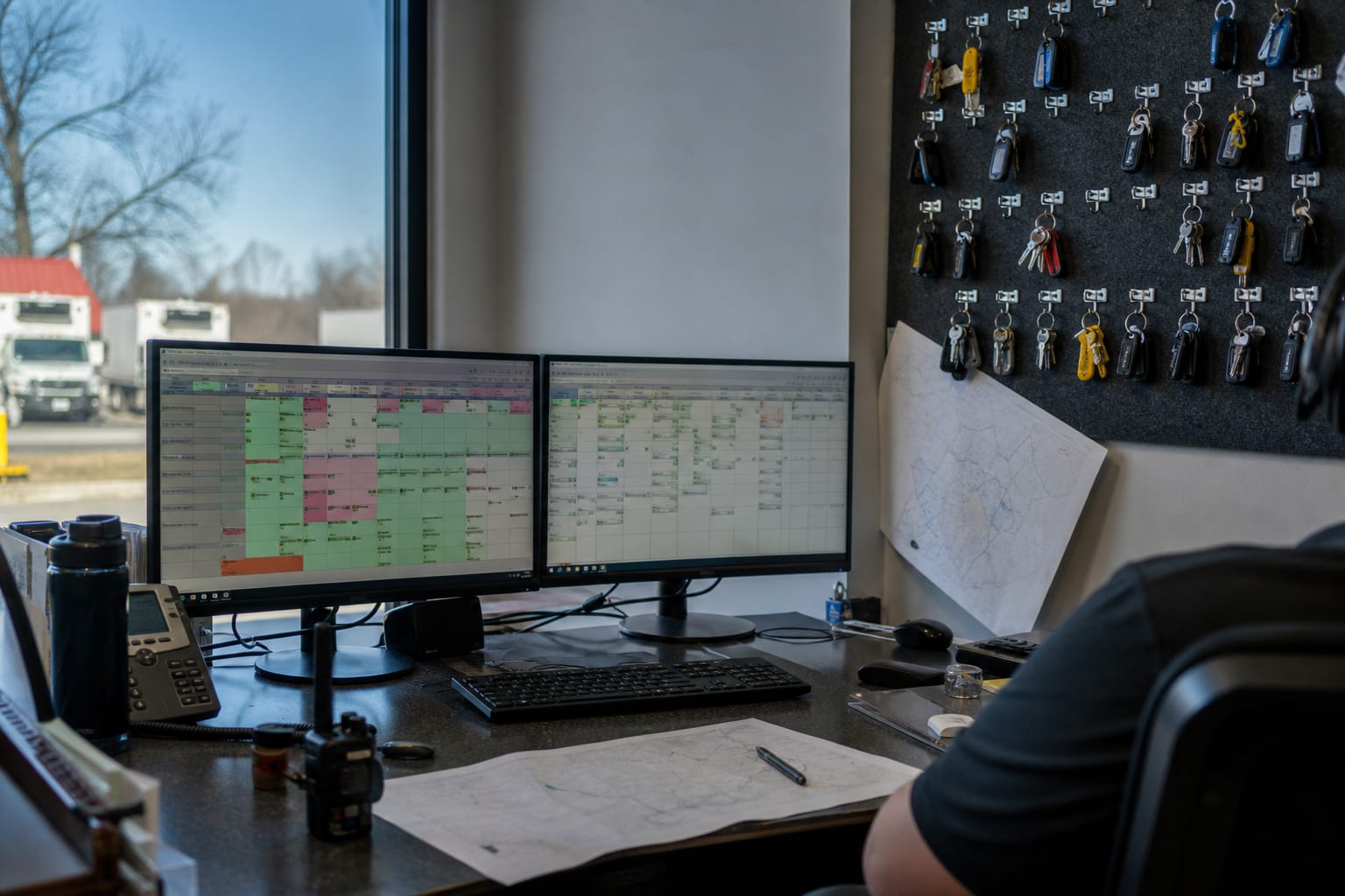 Dispatcher's dual-monitor desk showing crew assignments beside a truck-key board at dawn