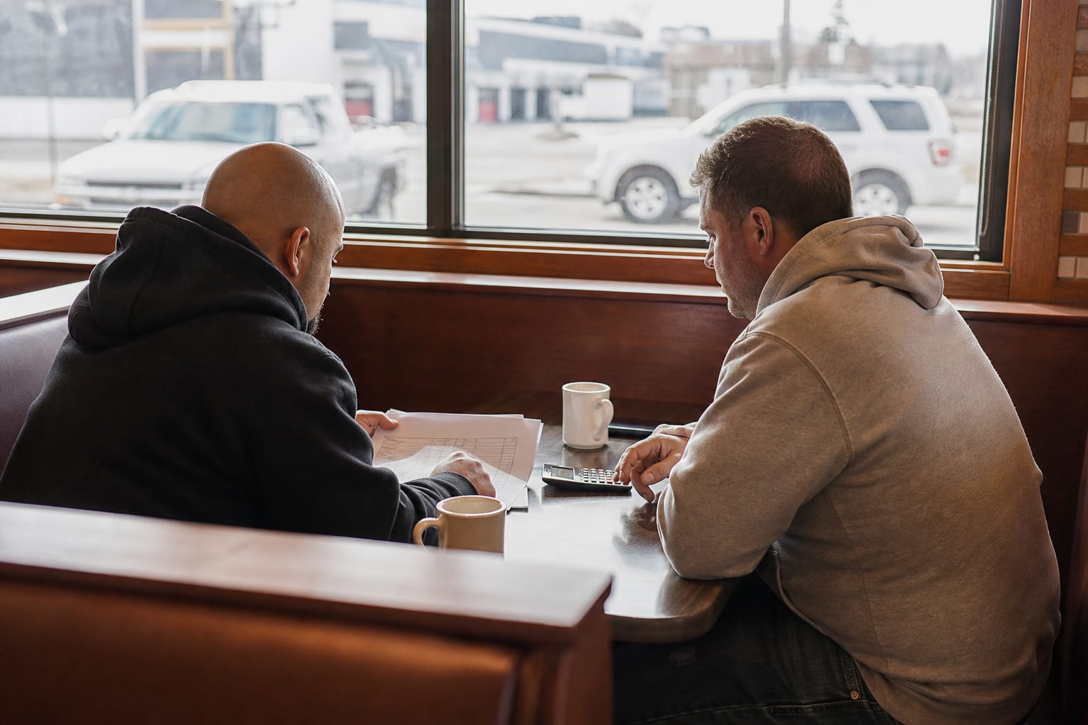 Two moving company owners in a diner booth reviewing a printed job-cost spreadsheet with a calculator