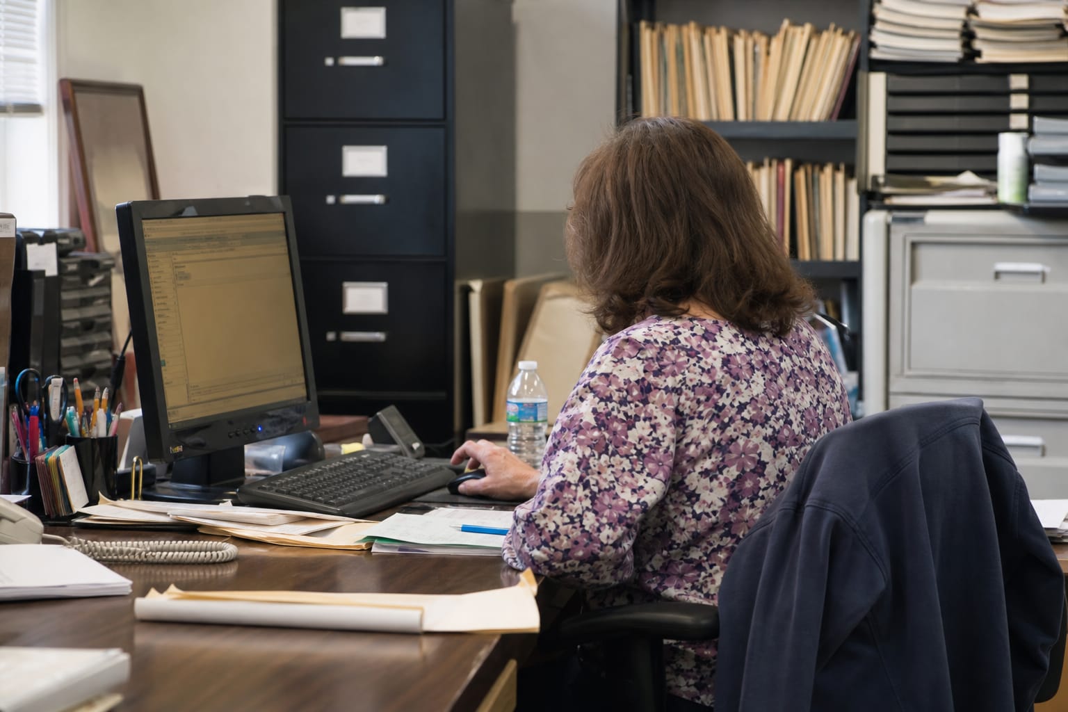 Office manager at an older desktop PC with paper move files and filing cabinets behind her.