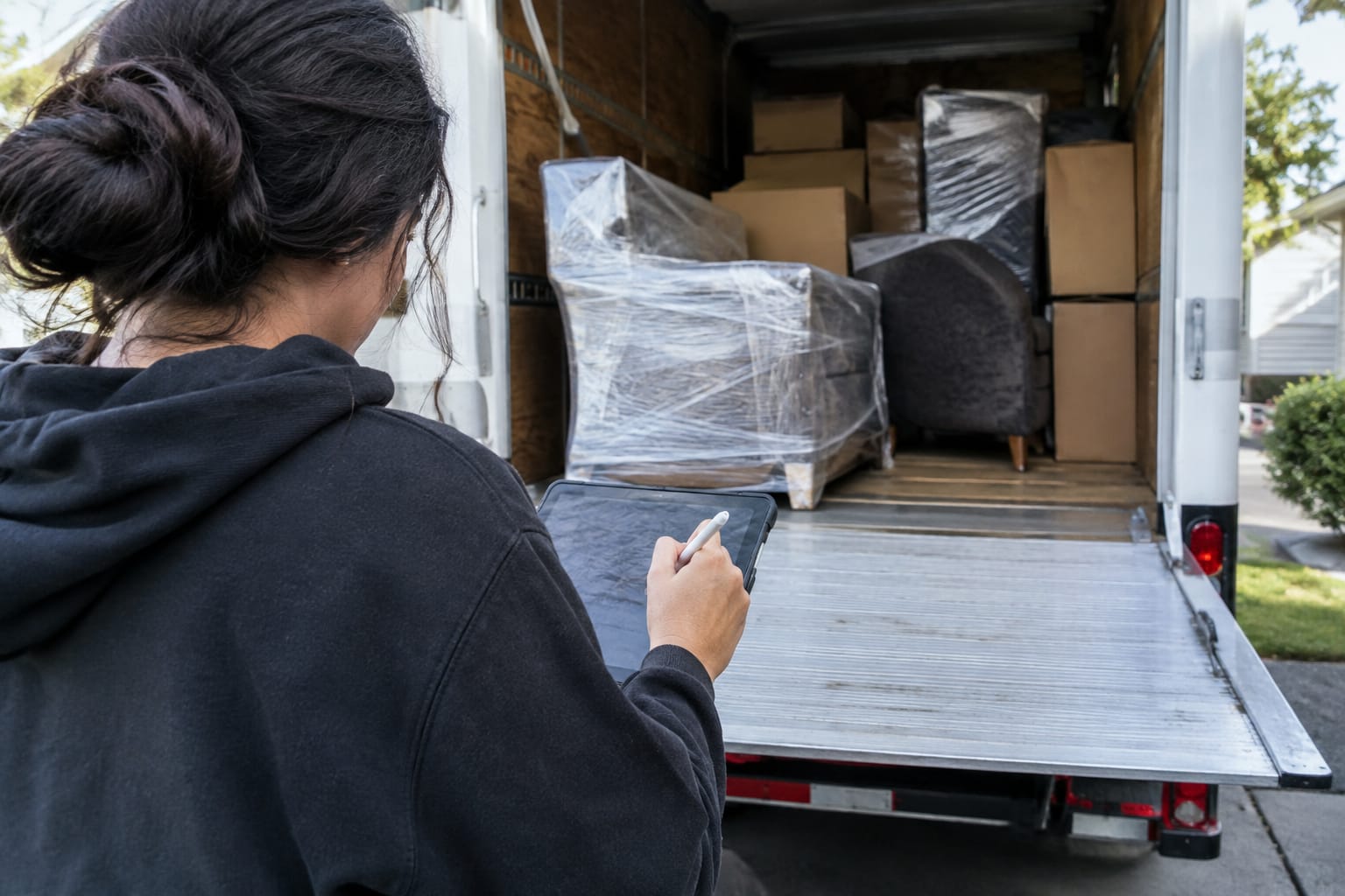 Customer signing a phone screen with a fingertip at a front door as a foreman holds it, moving truck at the curb