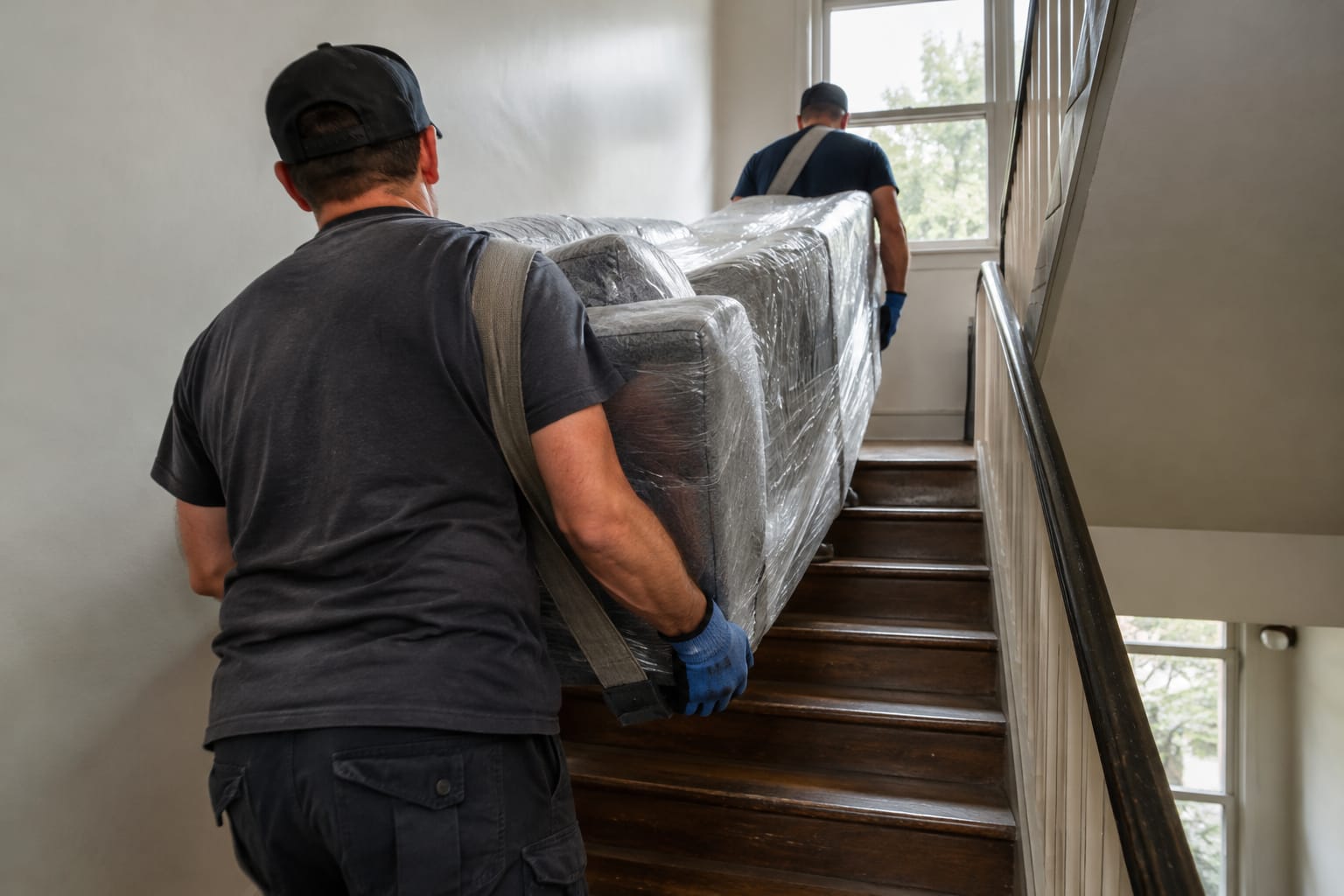 Two movers carrying a wrapped sofa up an outdoor apartment staircase, seen from the sidewalk