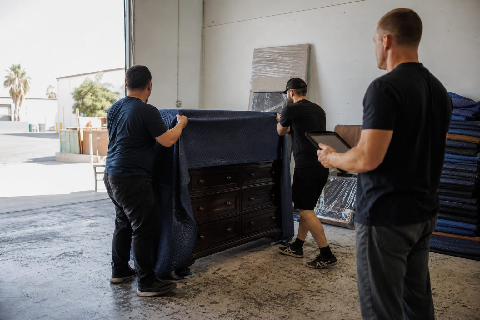 Two movers blanket-wrapping a dresser during warehouse training while a supervisor takes notes